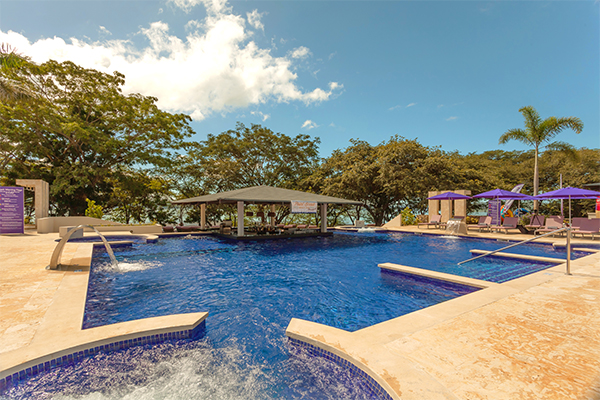 Large resort pool with blue water, sun loungers under purple umbrellas, thatched shelters, and tropical trees under clear sky at Planet Hollywood Costa Rica.