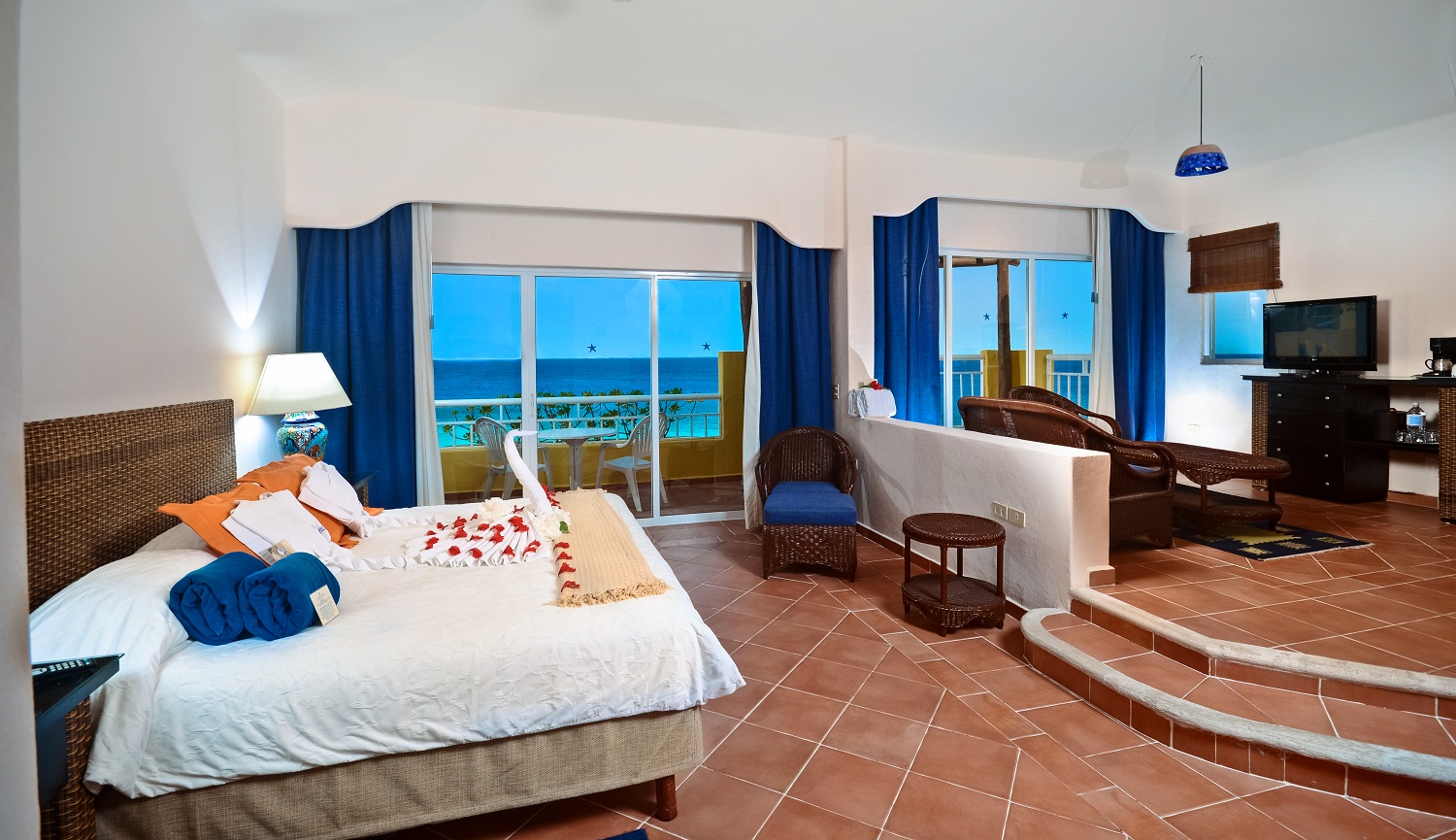 Oceanfront suite with king bed, blue curtains, terracotta tile floors, and balcony overlooking turquoise water at Playa Azul Cozumel.