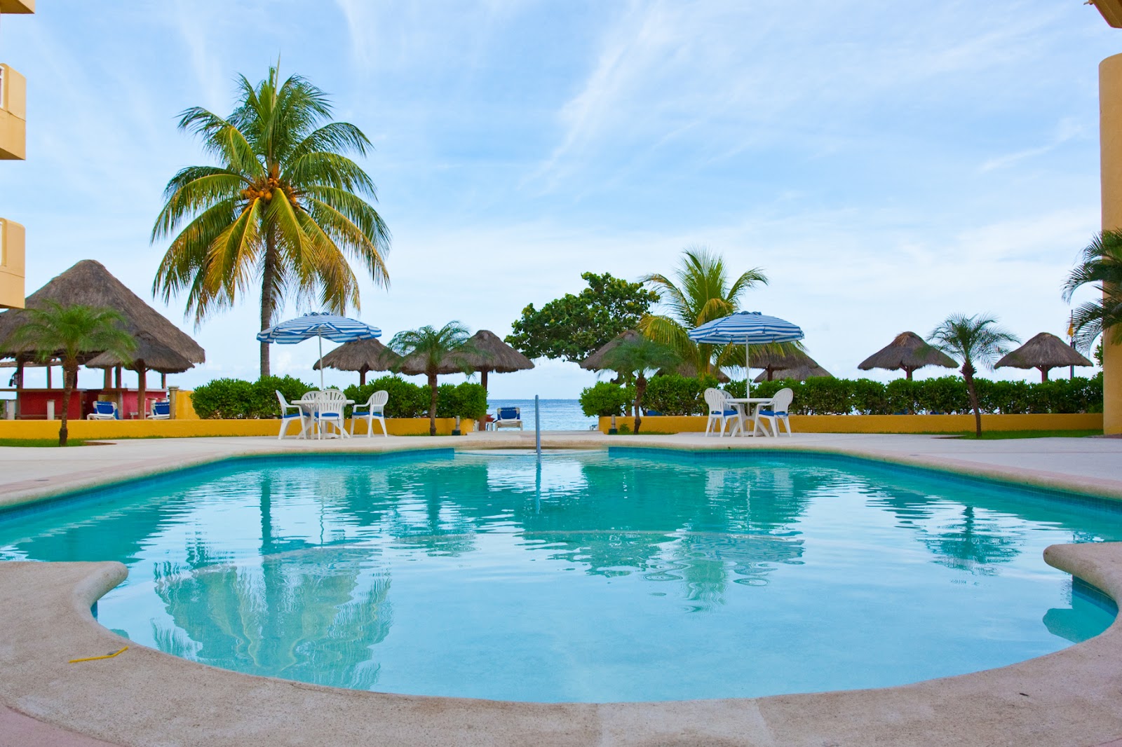 Resort pool with turquoise water, thatched umbrellas, white lounge chairs, and ocean view beyond yellow-trimmed deck.