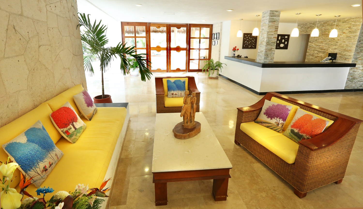 Resort lobby with yellow seating, wicker chairs, colorful pillows, potted palm, wooden sculpture, and reception desk with stone column.
