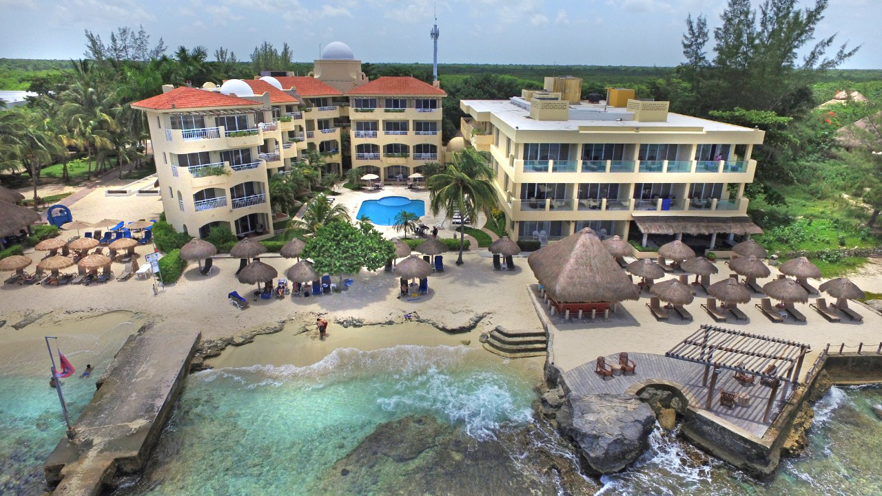 Playa Azul Cozumel Hotel's multi-story yellow buildings with red-roofed sections overlook a rocky beach with turquoise water, thatched umbrellas, and wooden docks.