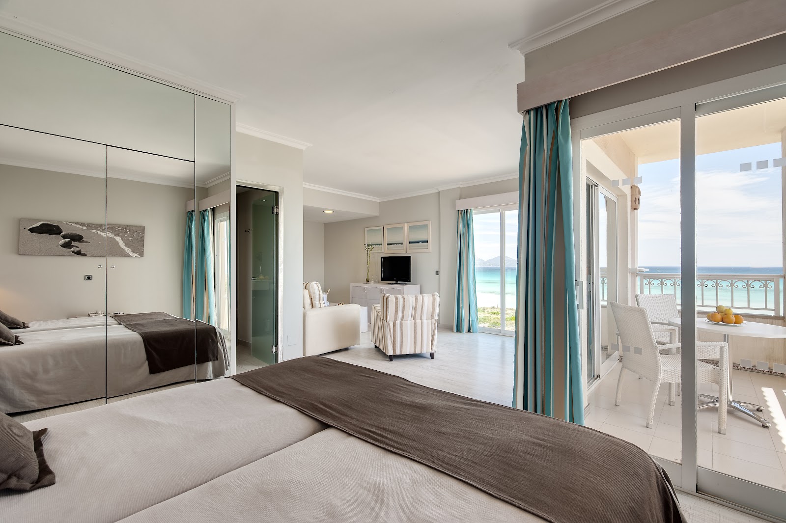 Bright beachfront suite with king bed, mirrored wardrobe, teal curtains, and ocean view from balcony at Playa Garden Selection Hotel & Spa.