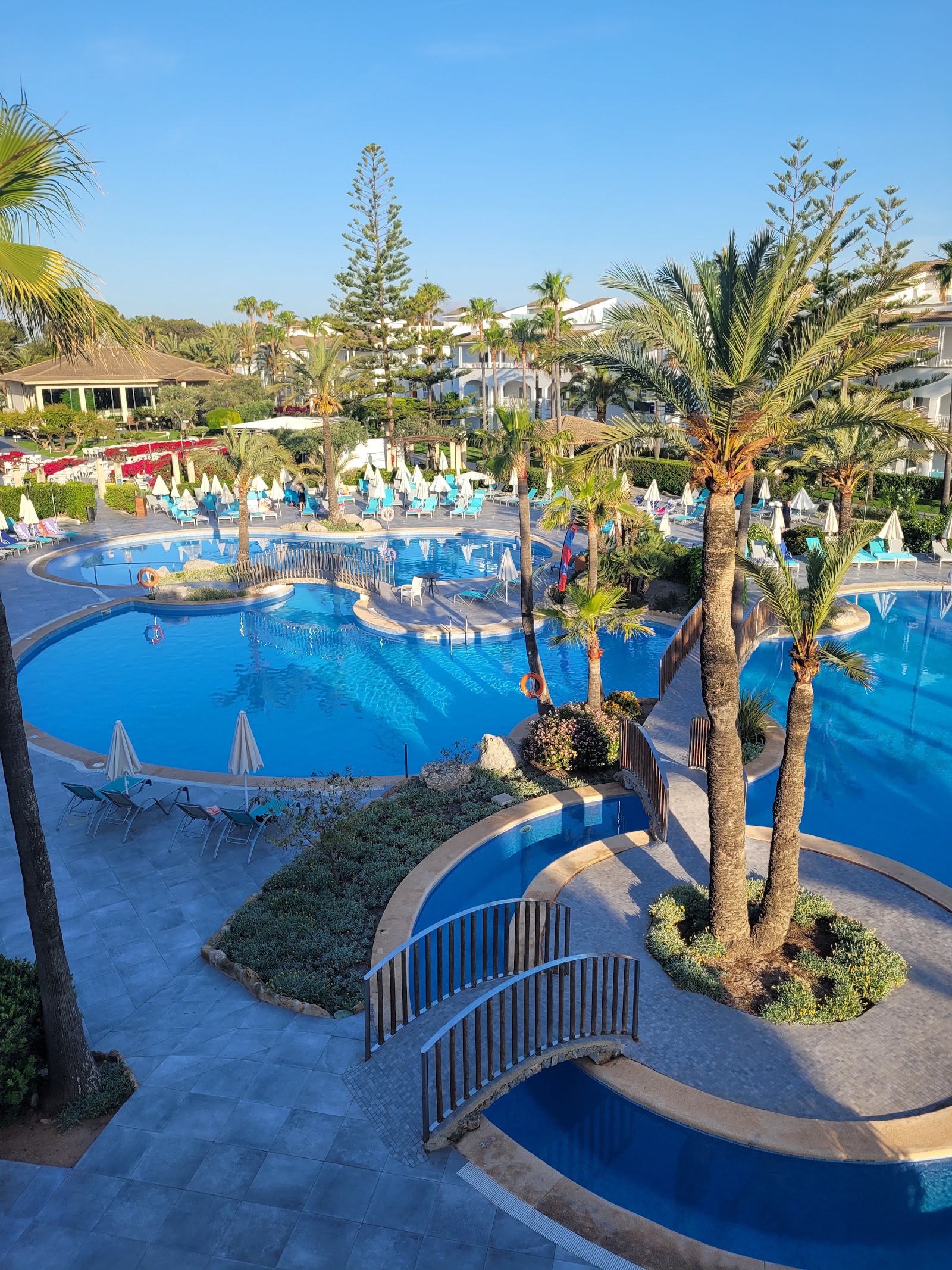 Blue pool with loungers in foreground, palm trees, white umbrellas, and multi-story resort buildings under clear sky at Playa Garden Selection Hotel & Spa.