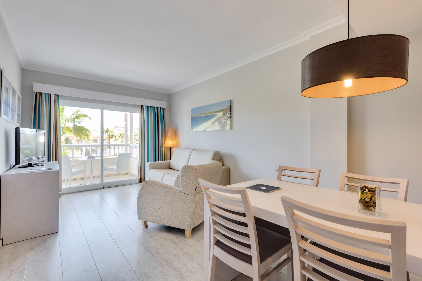 Resort room with dining table, upholstered chairs, sitting area, and balcony overlooking palm trees and ocean.