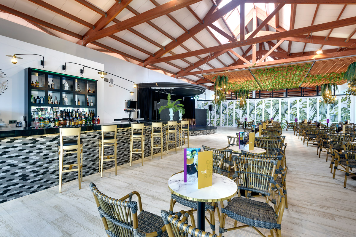 Modern resort bar and lounge with wooden beam ceiling, black and white tiled bar counter, woven rattan chairs, and tropical plant accents throughout the space.