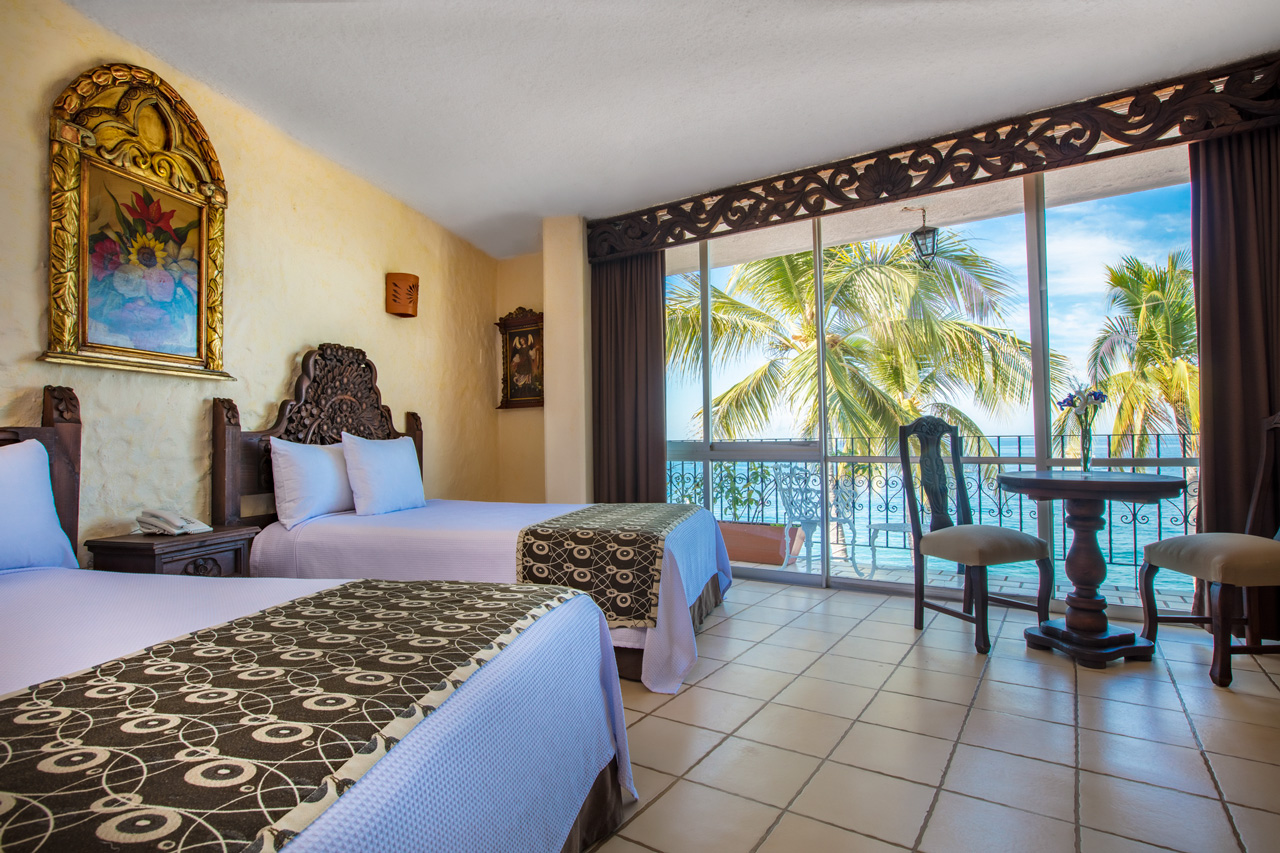 Hotel room with two beds, ornate wooden furniture, and ocean view balcony with palm trees at Playa Los Arcos.
