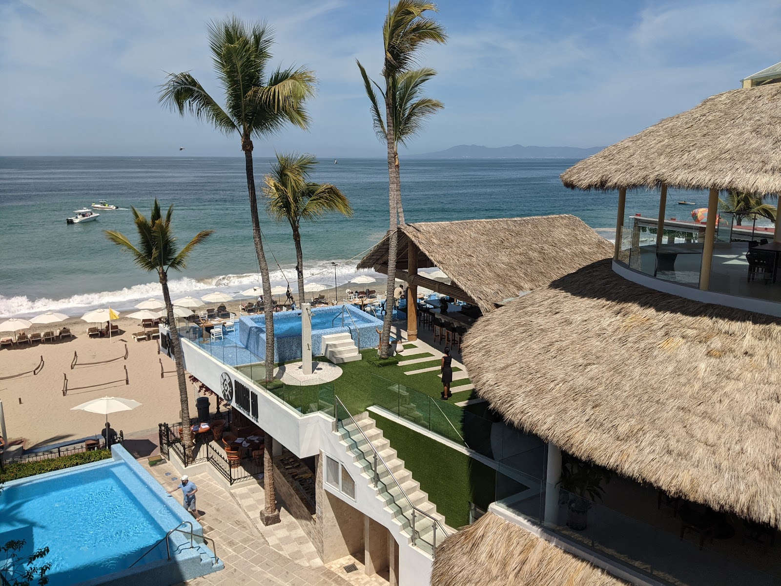 Playa Los Arcos resort overlooks a sandy beach with palm trees, thatched-roof structures, multiple pools, and the Pacific Ocean beyond.