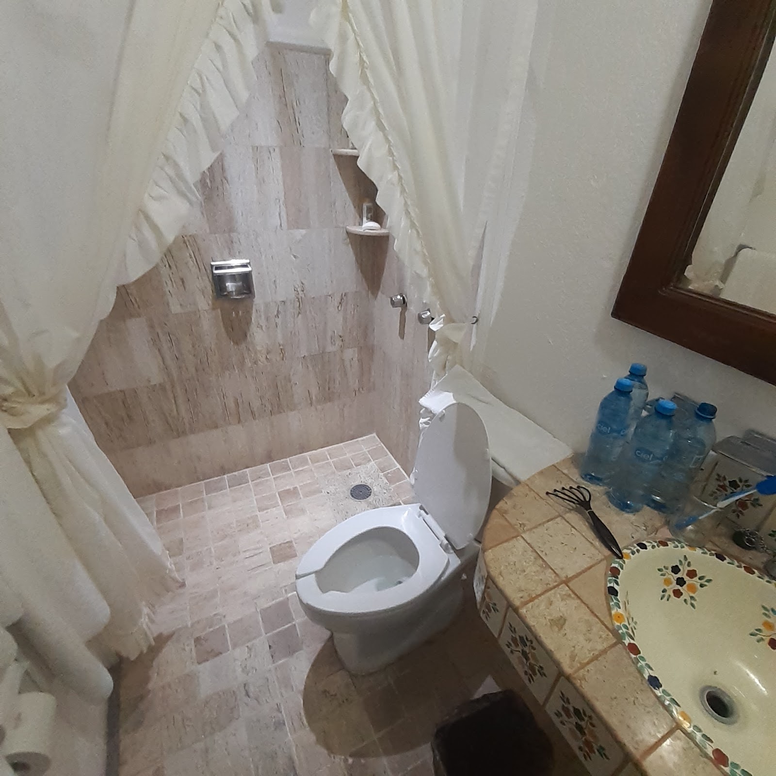 Bathroom with white toilet, walk-in shower with beige tile walls, sheer white curtains, water bottles on counter, and decorative sink area.