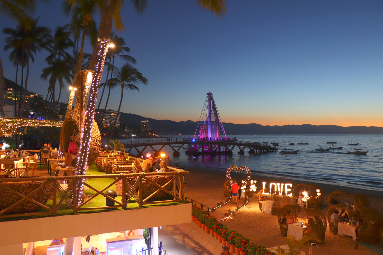 Beach venue at dusk with illuminated palm trees, dining areas, and a purple-lit pier extending into the ocean under a twilight sky.