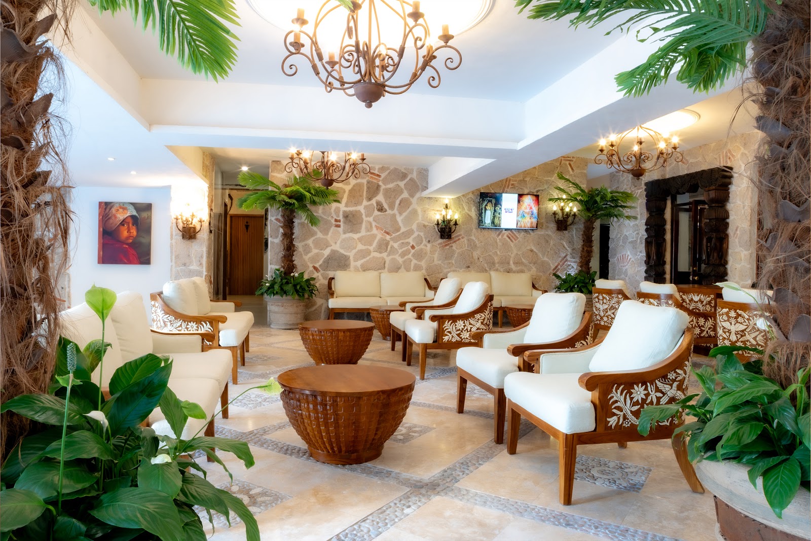 Indoor lounge at Playa Los Arcos with stone walls, carved wooden chairs with white cushions, woven side tables, hanging palms, and brass chandeliers.