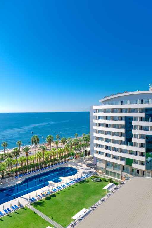 Modern waterfront resort with curved white building, blue pool, manicured lawn, palm trees, and Mediterranean Sea beyond under clear sky.