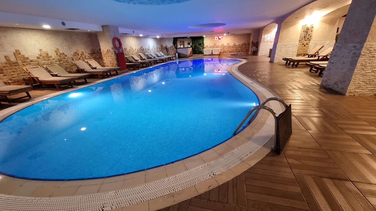 Indoor swimming pool at Porto Bello Hotel Resort & Spa with curved edge, metal ladder, wooden deck, and lounge seating along brick walls.