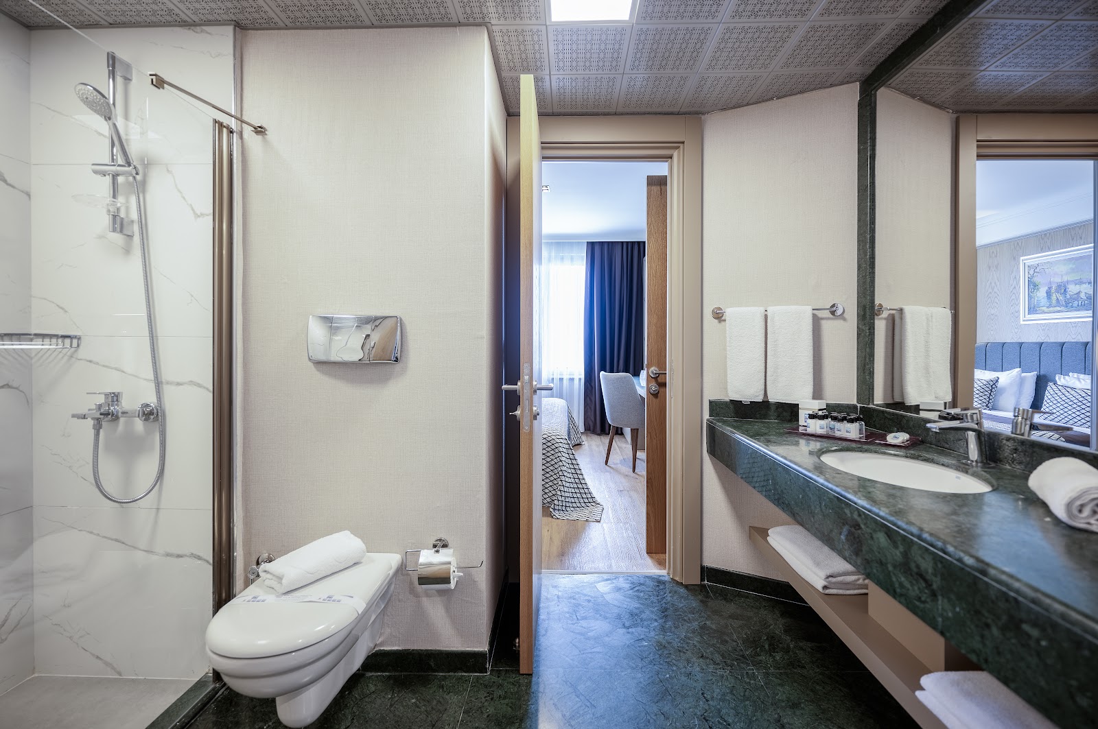 Modern bathroom with marble shower enclosure, dark green countertop sink, and open doorway revealing bedroom with ocean view at Porto Bello Hotel Resort & Spa.