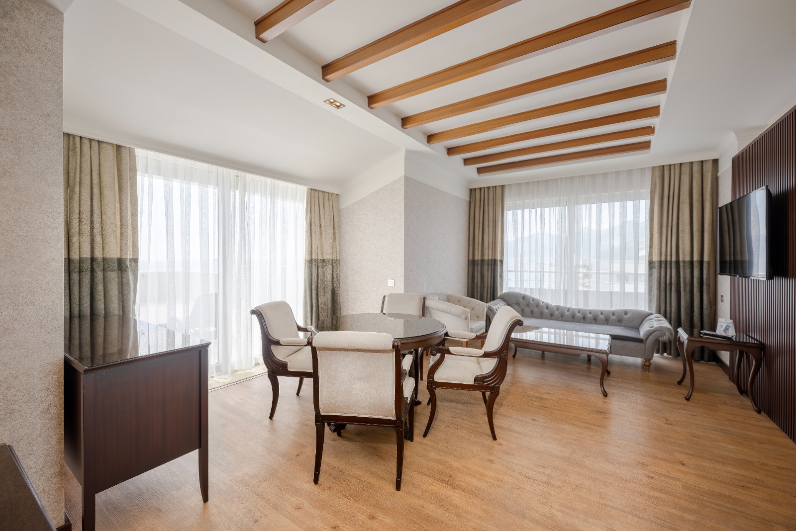 Spacious resort suite with wooden beams, cream upholstered furniture, and floor-to-ceiling windows overlooking the coast.