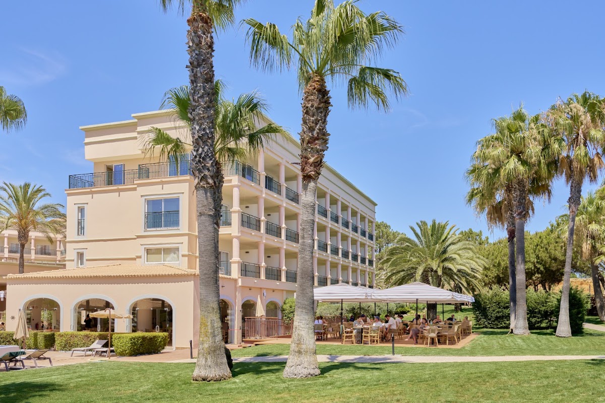 Beige resort building with palm trees, arched ground-floor colonnade, and outdoor dining area under white umbrellas on manicured lawn.