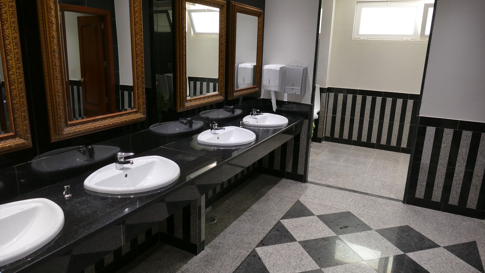 Public restroom at PortoBay Blue Ocean with multiple white sinks, gold-framed mirrors, black countertop, and diamond-patterned floor tiles.