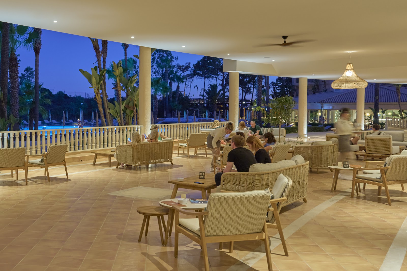Poolside lounge area with rows of sunbeds and dark umbrellas overlooking multiple pools, palm trees, and the sea at PortoBay Blue Ocean.