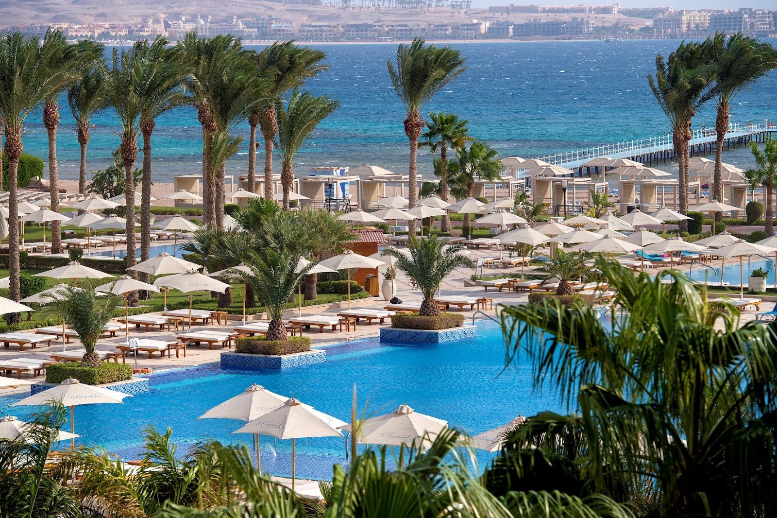 Resort pool with white umbrellas and loungers surrounded by palm trees, beach cabanas, and Mediterranean Sea beyond.