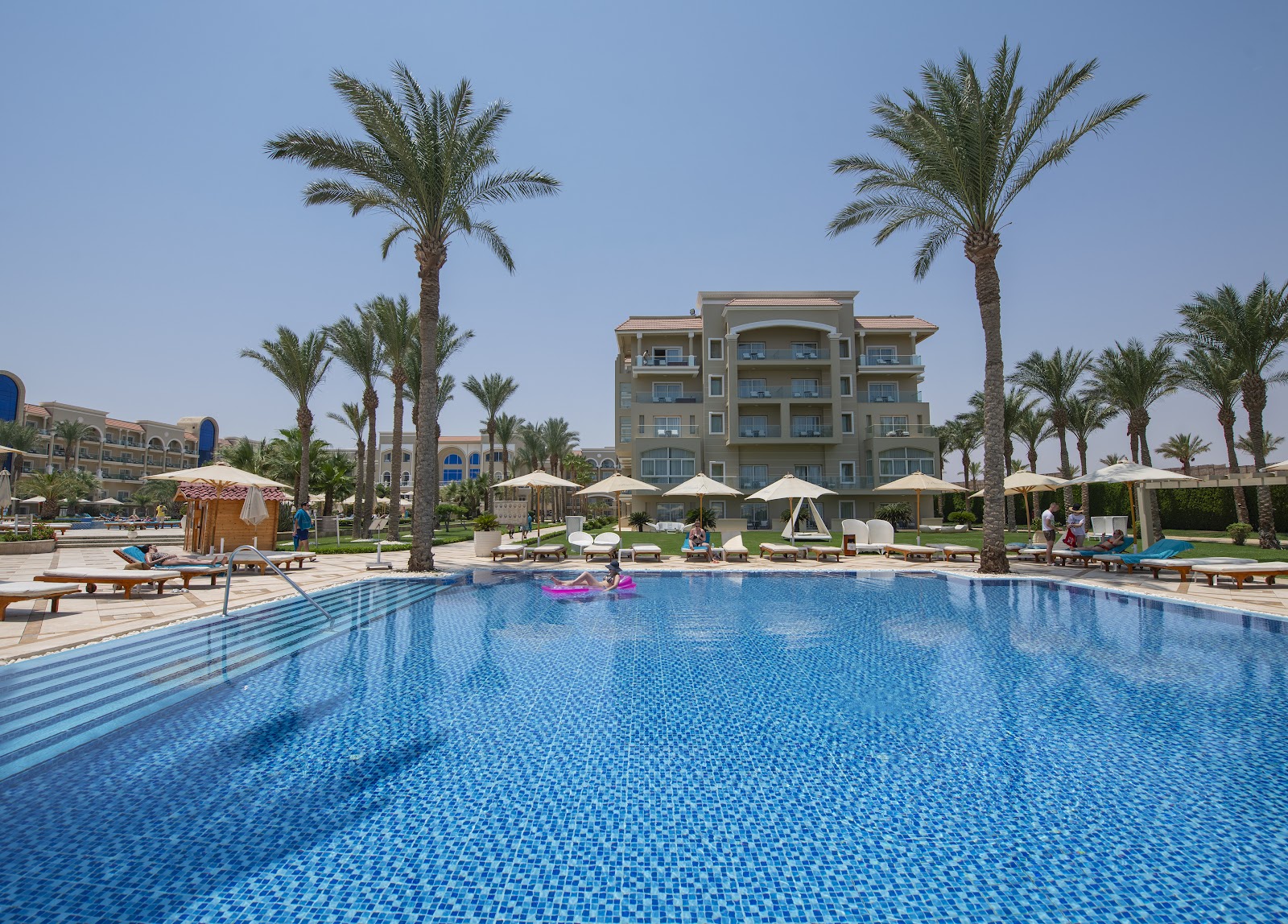 Large swimming pool with guests on loungers and umbrellas, palm trees, and multi-story resort buildings under clear sky.