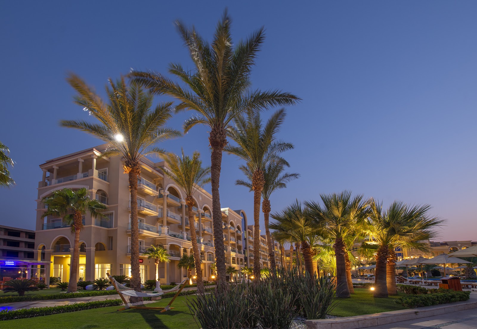 Palm trees and landscaped grounds frame a beige resort building illuminated at dusk, with arched windows and multiple stories visible.
