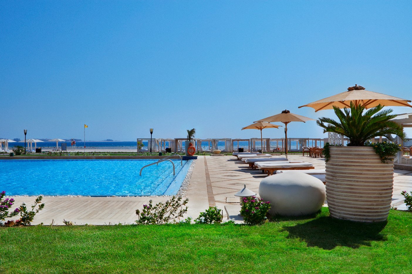 Pool deck at Premier Le Reve Hotel & Spa with loungers, tan umbrellas, and sea view beyond the beachfront fence.