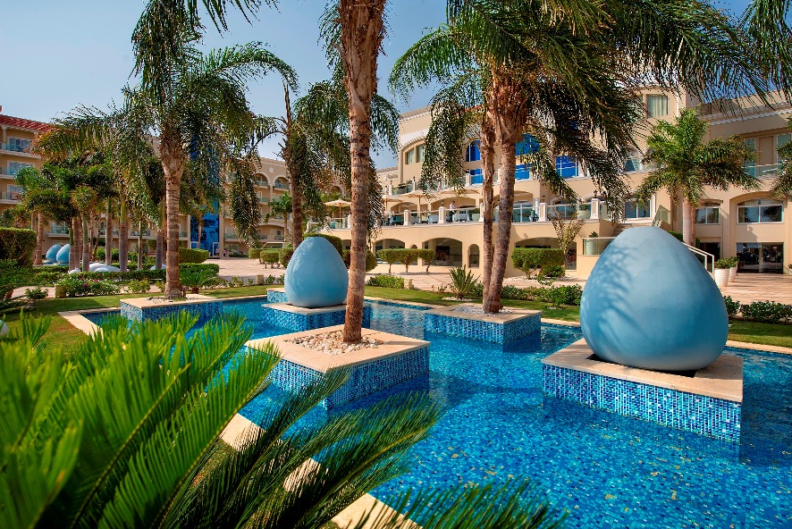 Resort courtyard with blue-tiled pools, tall palm trees, large blue sculptural spheres, and tan multi-story hotel buildings under clear sky.