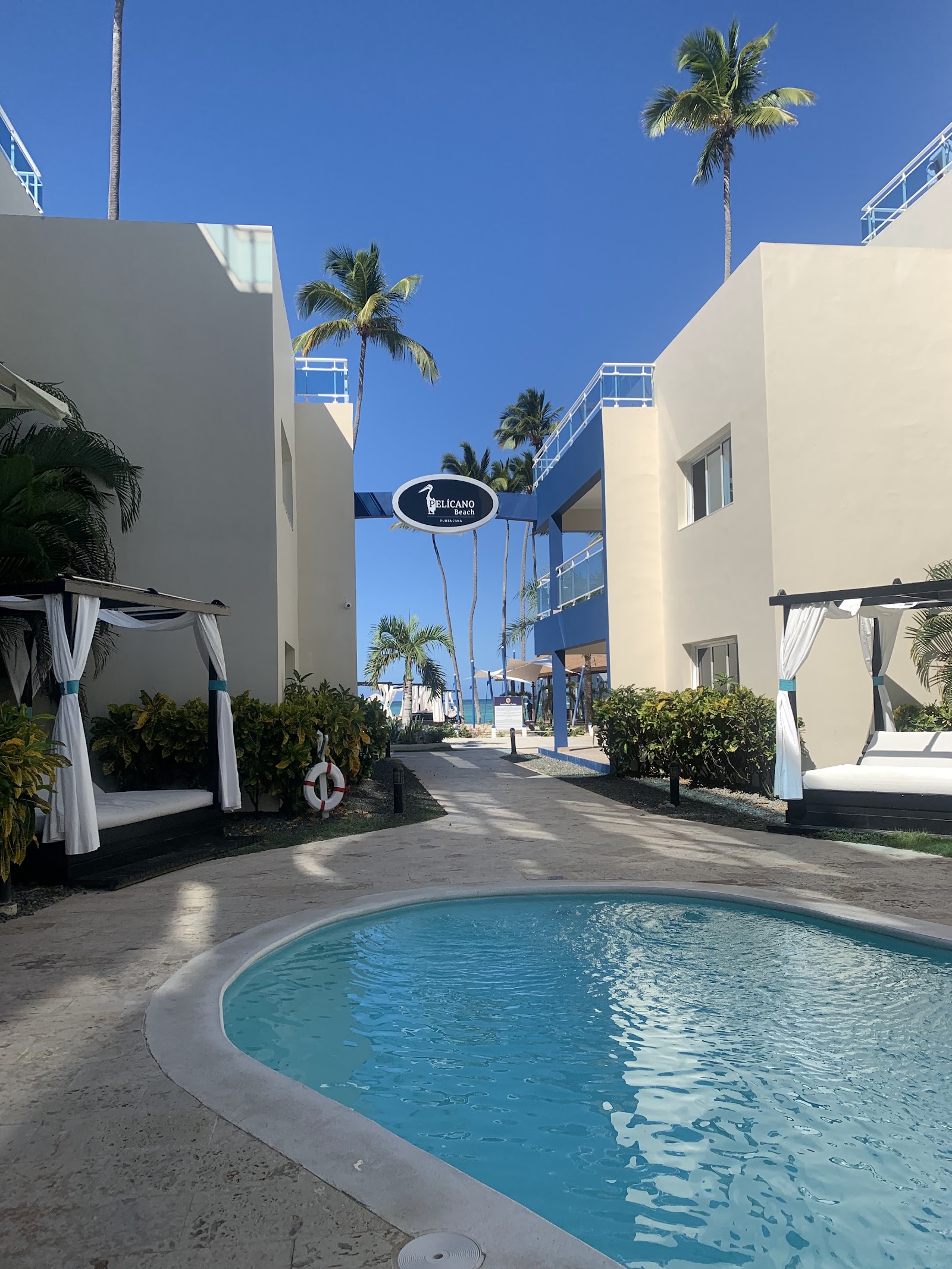 Pelicano Resort courtyard with round pool, white buildings, palm trees, and blue archway under clear sky.
