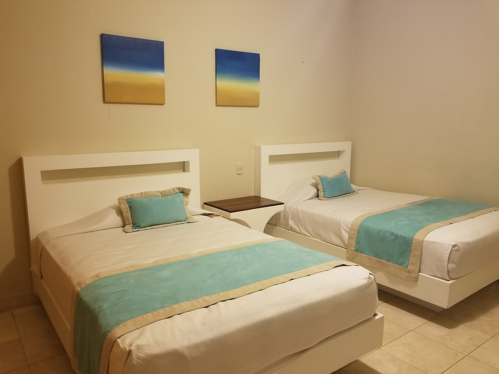 Double room with two beds featuring white headboards, turquoise runners, and matching throw pillows in a cream-colored room with landscape artwork.