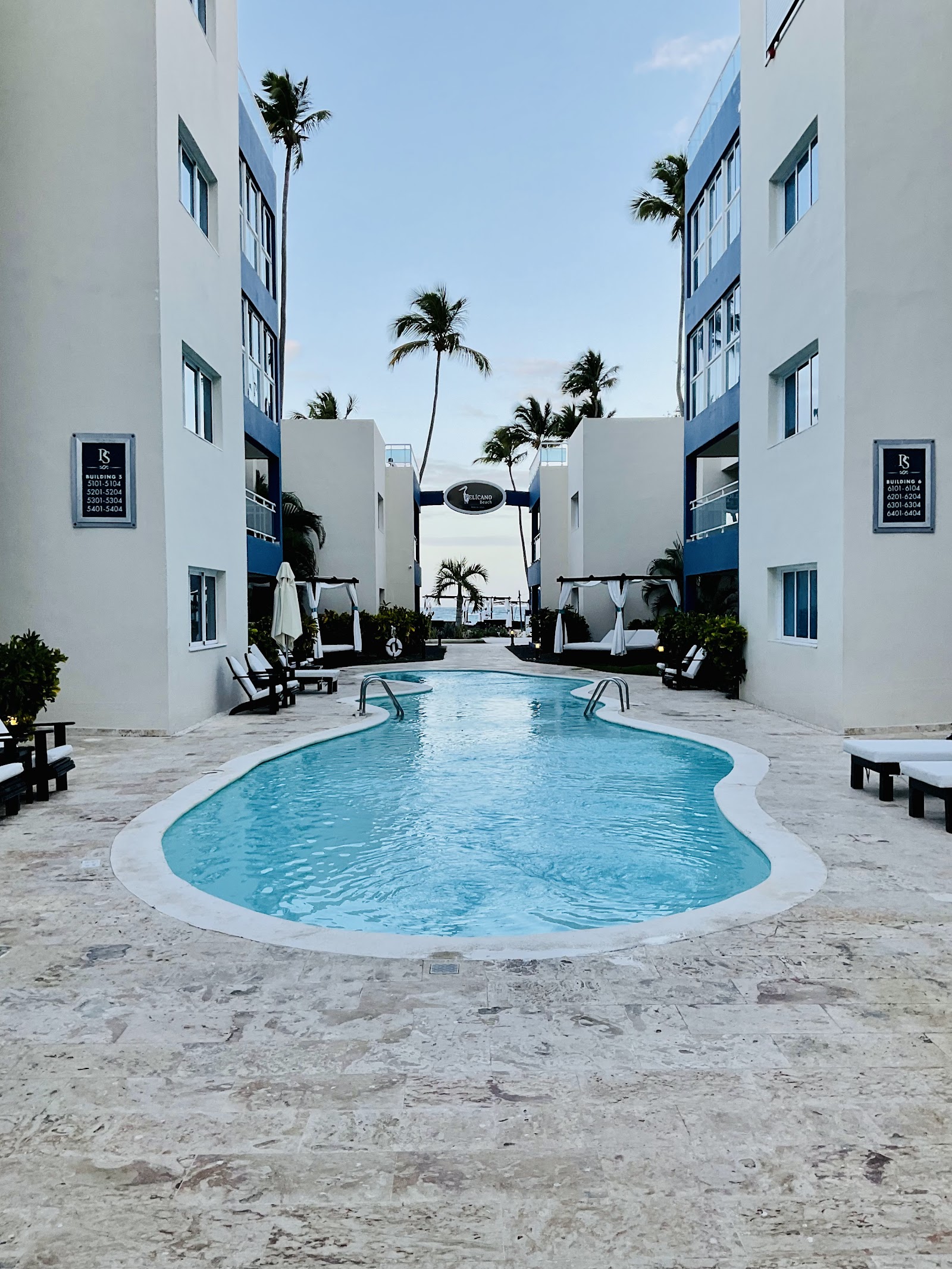 Curved pool flanked by white resort buildings with blue trim, palm trees, and lounge chairs in a courtyard leading to the ocean.