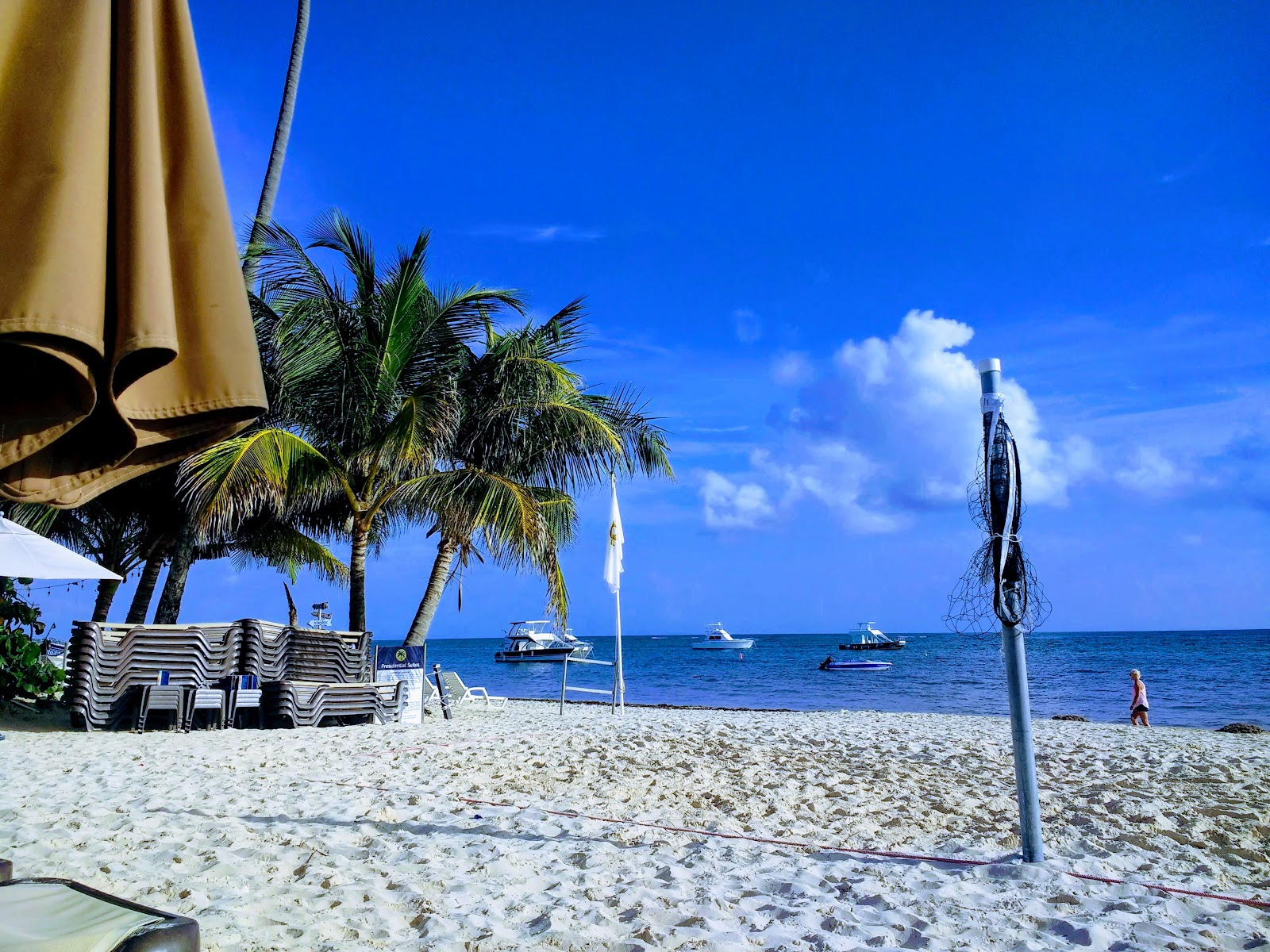 Sandy beach with lounge chairs and tan umbrellas under palm trees, sailboats anchored in clear blue water.