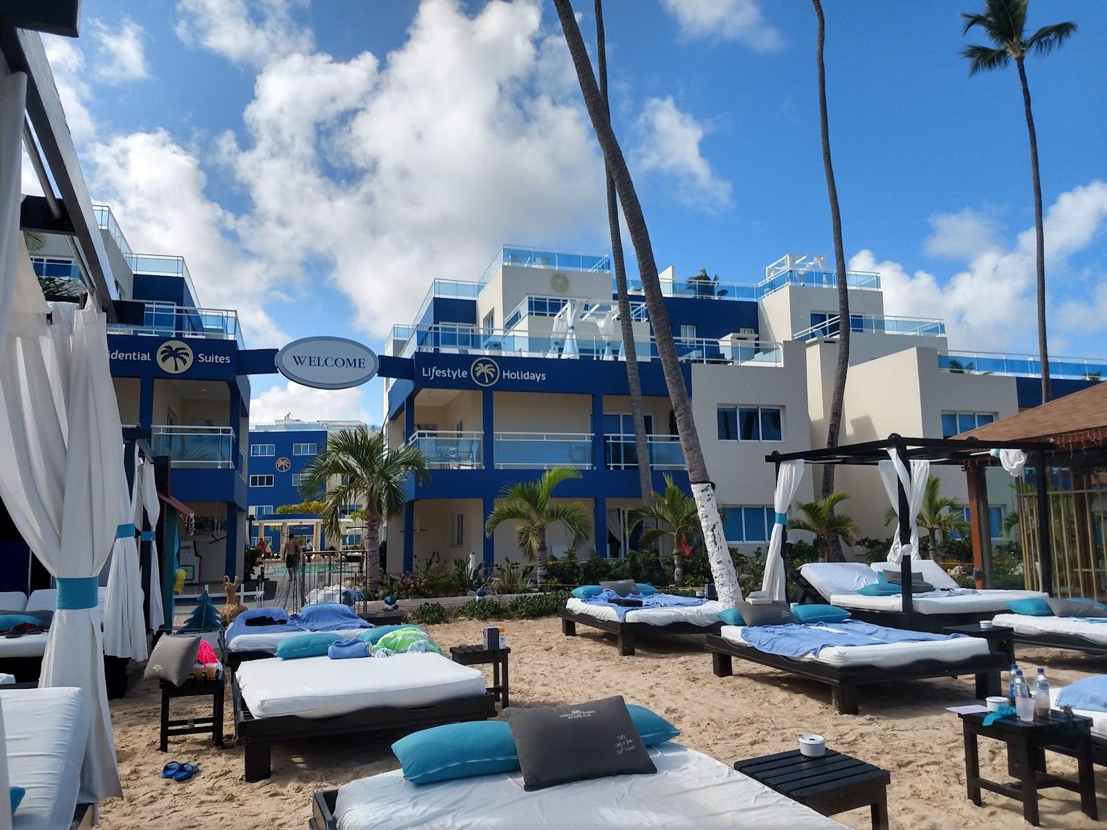 Beach lounge area at Presidential Suites with white and blue cushioned sun beds, palm trees, and the blue and cream resort building behind.