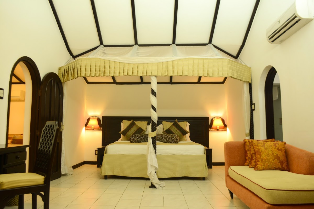 Resort bedroom with canopy bed draped in cream fabric, black wooden frame, patterned pillows, wall sconces, orange sofa, and arched doorways.