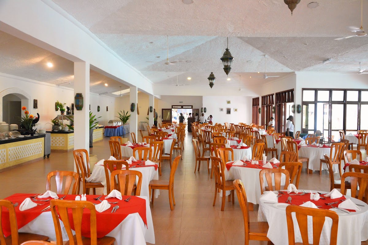 Spacious dining room with wooden chairs at tables set with red and white linens, hanging lanterns, and open doors leading to outdoor areas.