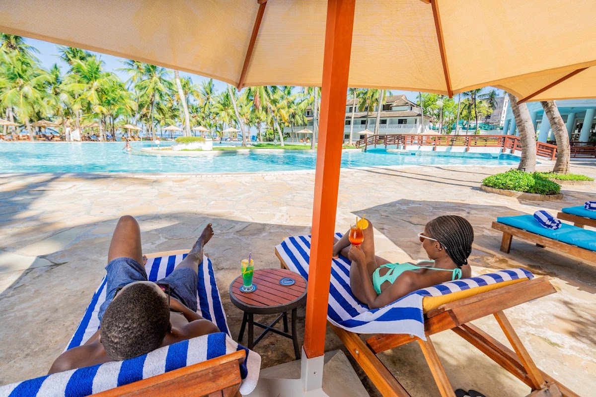 Guests relax on striped lounge chairs under an orange umbrella by the pool at PrideInn Flamingo Beach Resort & Spa, with palm trees and resort buildings visible beyond.