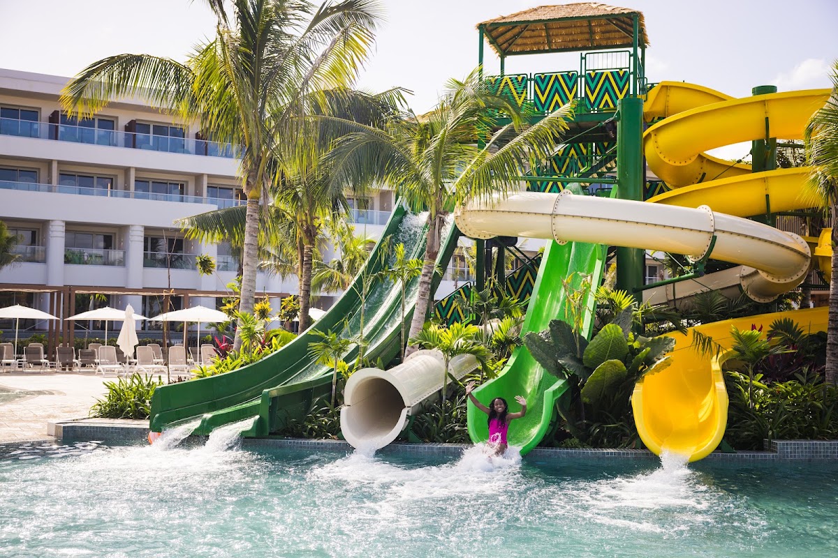 Water slides with green and yellow tubes flow into a shallow pool where a child plays, with the white resort building and palm trees behind.