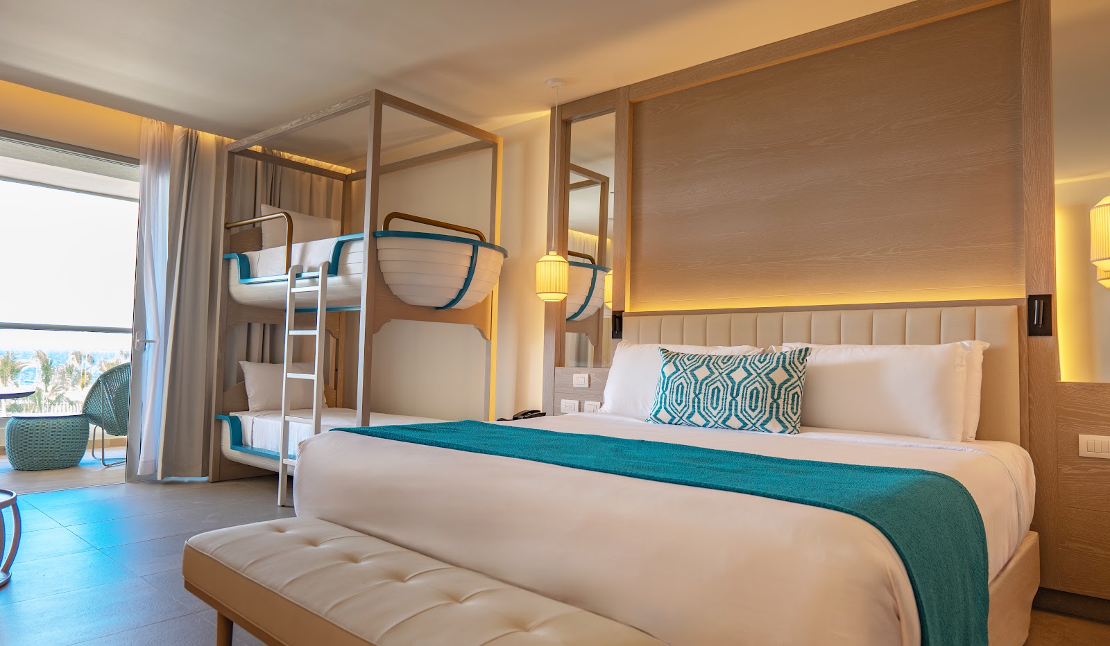 Resort suite with queen bed, bunk beds, teal accents, warm lighting, and ocean view through floor-to-ceiling windows.