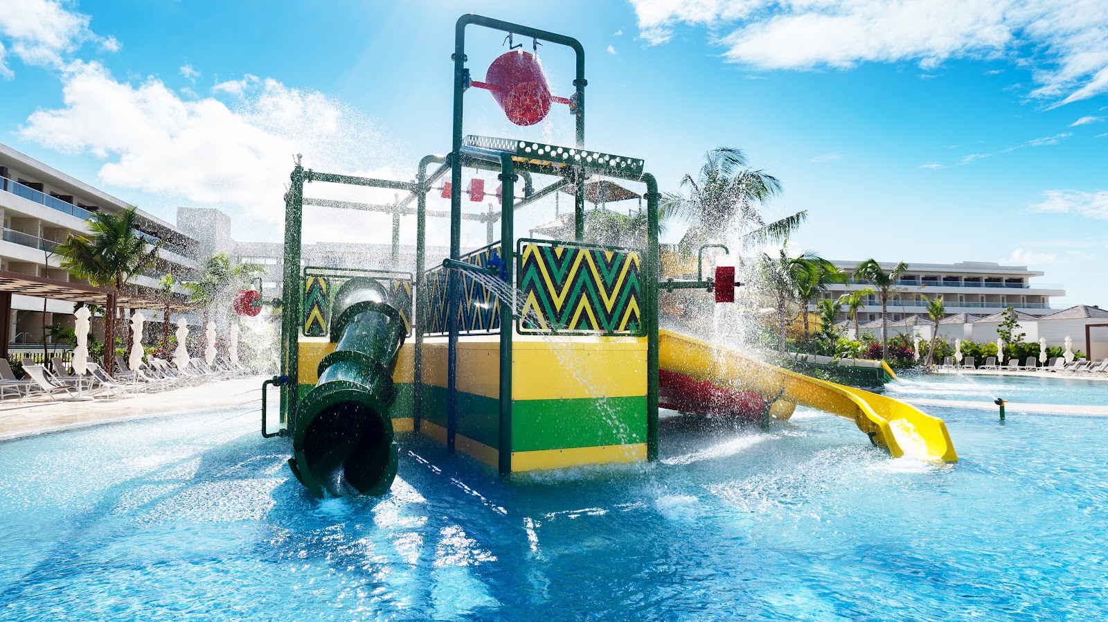 Colorful water play structure with slide and bucket dumper in shallow pool at Princess Grand Jamaica, flanked by resort buildings and palm trees.