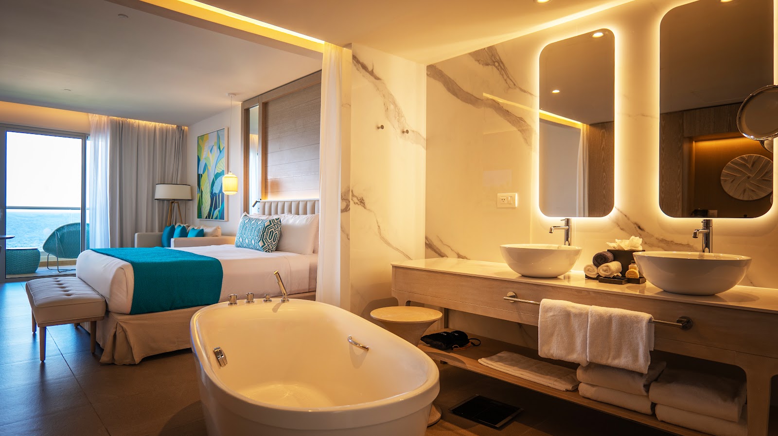 Resort suite with freestanding bathtub in foreground, dual vanity with illuminated mirrors, and bedroom area with desk visible beyond.