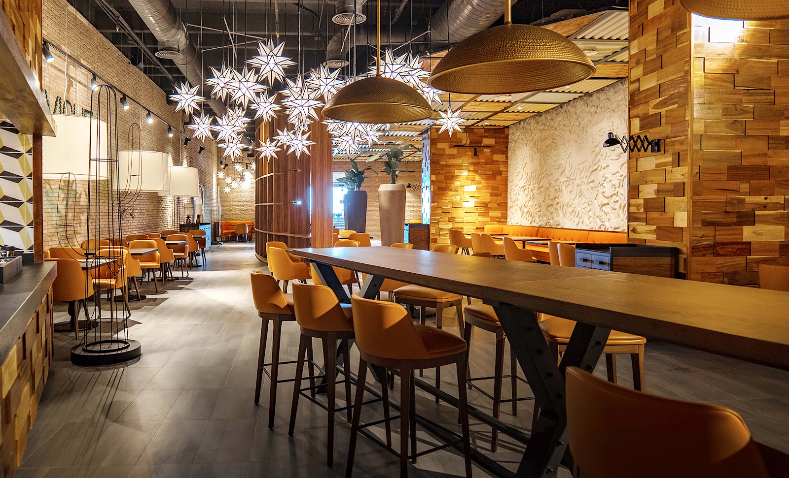 Modern resort lounge with wooden accent walls, orange chairs at high-top tables, and illuminated star-shaped pendant lights hanging from industrial ceiling.