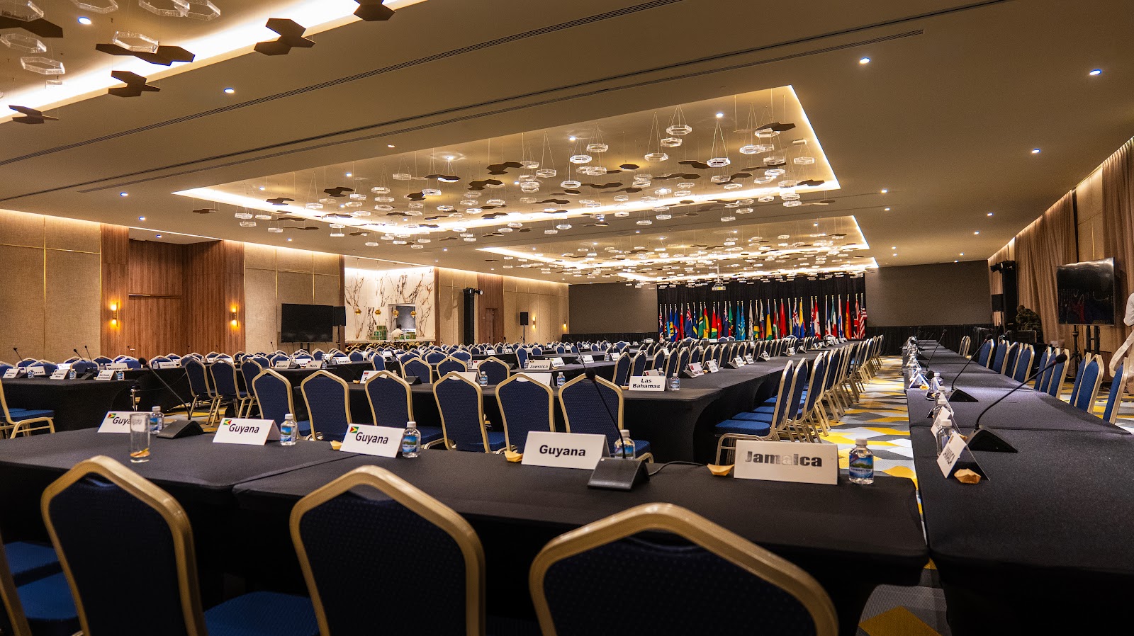 Large conference ballroom with rows of tables, blue and gold chairs, country name placards, and flags displayed along the back wall under recessed lighting.