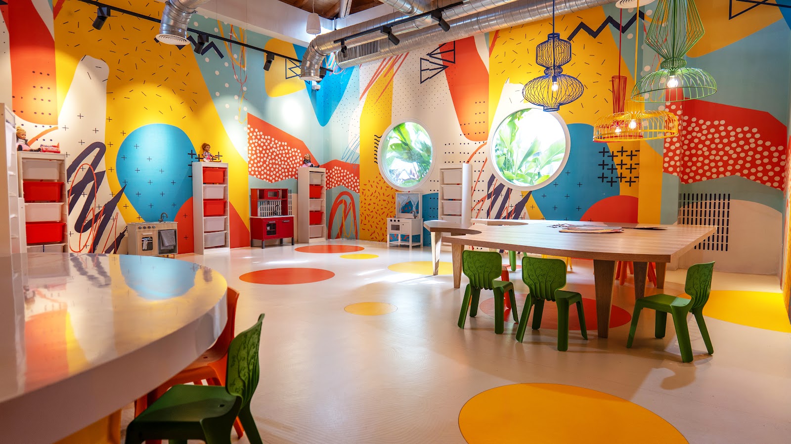 Colorful kids' club room with yellow, red, and blue walls, green chairs, wooden table, hanging lanterns, and circular floor decorations.
