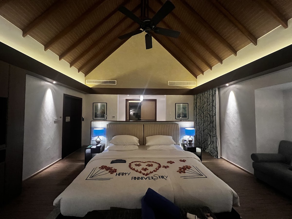 Resort suite with king bed decorated with Happy Anniversary petals, vaulted wood ceiling with ceiling fan, and blue-lit nightstands on either side.