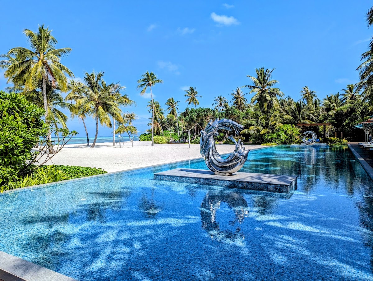Infinity pool with spiral sculpture overlooking white sand beach lined with palm trees under clear blue sky at Pullman Maldives Maamutaa.