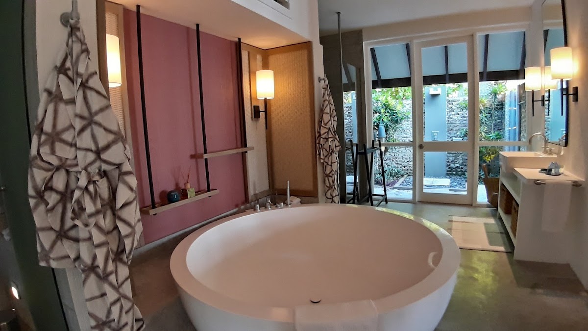 Spacious resort bathroom with round soaking tub, pink accent wall, vanity sink, patterned towels, and glass doors opening to tropical garden courtyard.