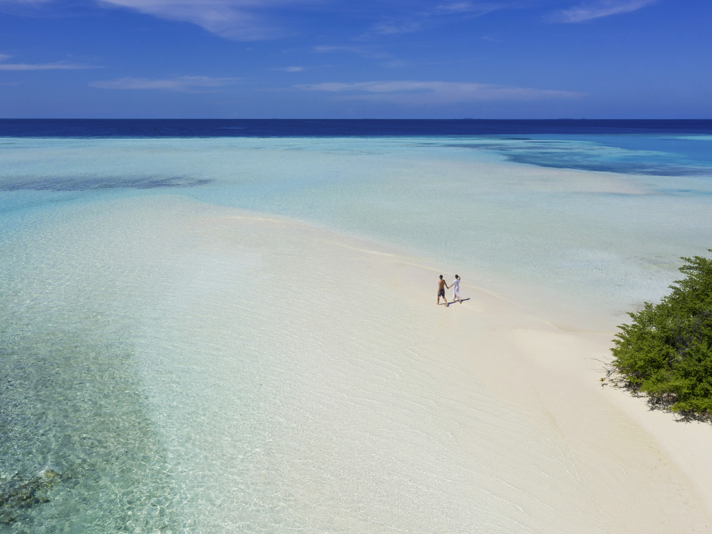 Two guests walk in shallow turquoise water at Pullman Maldives Maamutaa with white sand beach and mangroves visible.