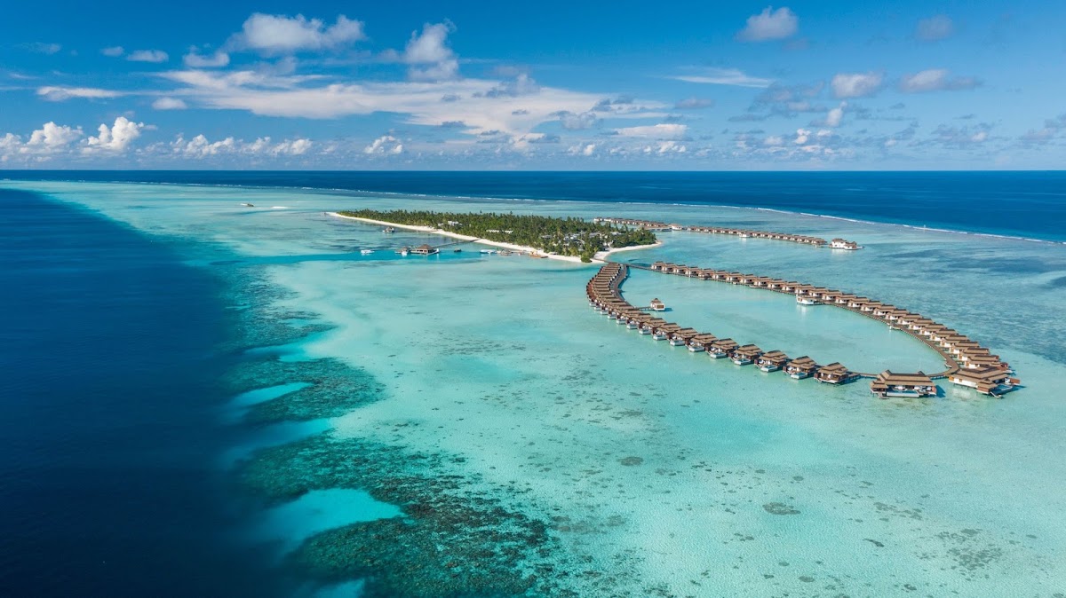 Aerial view of Pullman Maldives Maamutaa with curved rows of overwater bungalows in turquoise lagoon, palm-lined island, and deep blue ocean beyond.