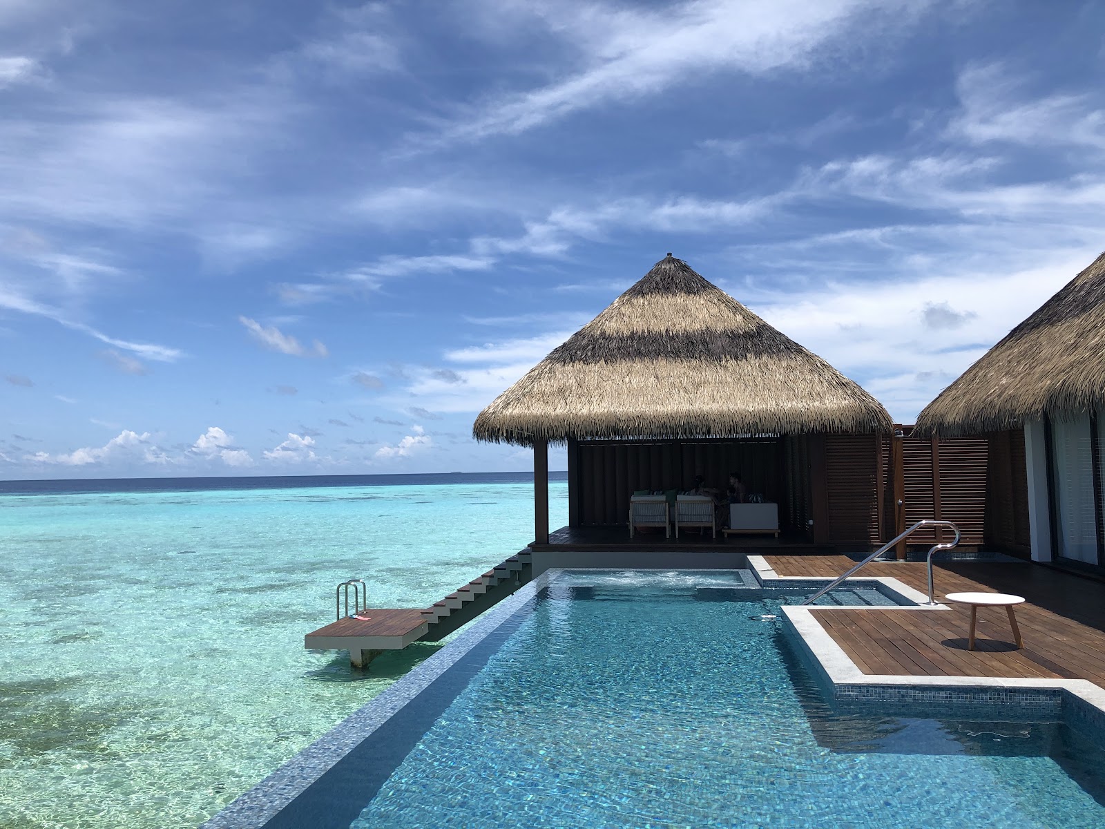 Wooden deck with blue lounge chair and infinity pool overlooking turquoise ocean under bright sun and blue sky at Pullman Maldives Maamutaa.