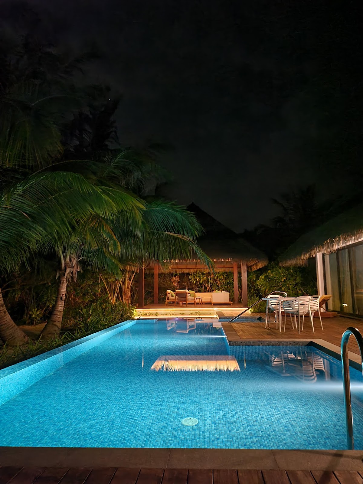 Illuminated infinity pool with thatched-roof bungalow and dining pavilion at night, palm trees and wooden deck surrounding the water.