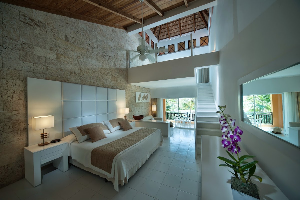 Modern resort bedroom with stone walls, king bed, purple orchid on windowsill, and ocean-view balcony visible through glass doors.