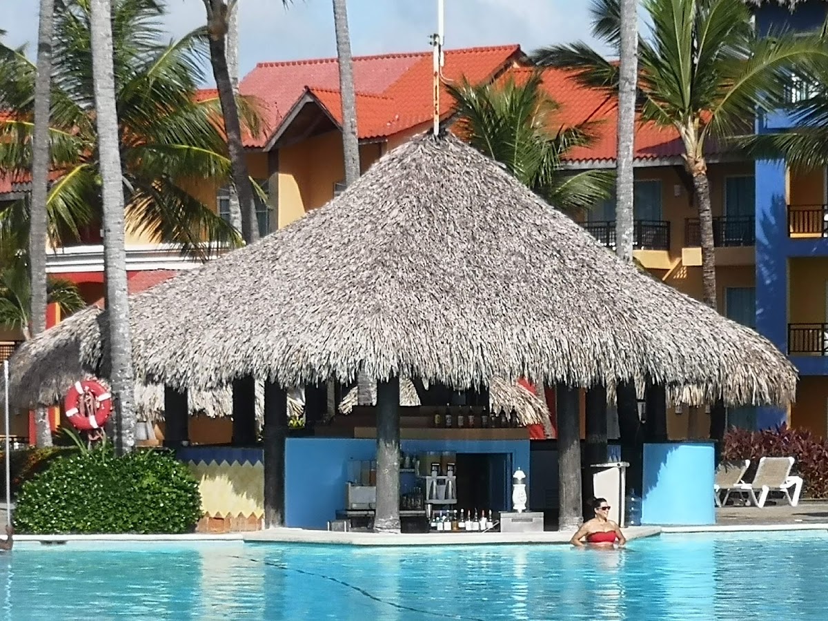 Thatched-roof palapa with blue base by the pool at Punta Cana Princess, palm trees and colorful colonial buildings behind.
